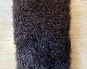May include: A brown fur boot cover with a leather strap and buckle at the top. The boot cover has a zipper at the bottom. The fur is dark brown and covers the majority of the boot cover.