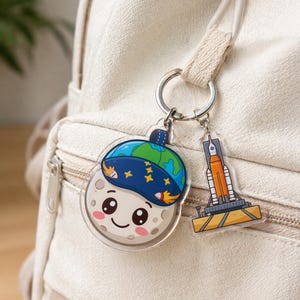 May include: A clear acrylic keychain featuring a smiling moon character wearing a blue and green Earth-themed hat, and a rocket ship. The keychain is attached to a silver keyring, perfect for keys or bags.