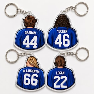 May include: Four clear acrylic keychains featuring the backs of people wearing blue jerseys with white numbers and names. The names are Graham, Tucker, Di Laurentiis, and Logan. Each keychain has a silver metal ring.