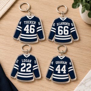 May include: Four acrylic hockey jersey keychains. Each keychain is navy blue with white stripes and lettering. The jerseys display the names "Tucker", "Di Laurentis", "Logan", and "Graham" with the numbers 46, 66, 22, and 44.