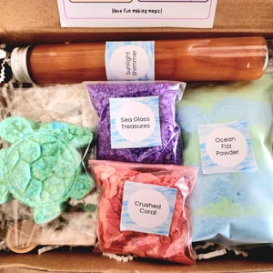 May include: A craft kit in a cardboard box containing a turtle-shaped bath bomb, a tube labelled Sunlight Shimmer, a bag of purple Sea Glass Treasures, a bag of pink Crushed Coral, and a bag of blue and green Ocean Fizz Powder with a label.