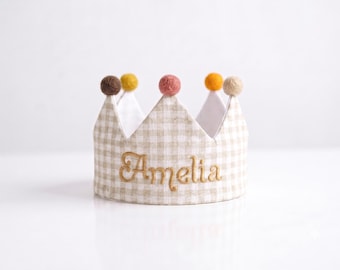 May include: A beige and white gingham fabric crown featuring the embroidered name Amelia in gold thread. The crown is topped with five small, round, felt pom-poms in shades of brown, yellow, pink, orange, and cream, with a white interior lining.