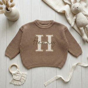 May include: A brown knit sweater with the name "Henry" embroidered on the front. The letter "H" is in a light beige gingham pattern, and the name is in a cursive font. The sweater has long sleeves and a crew neck.