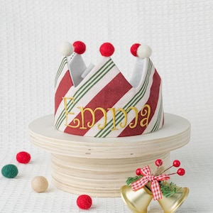 Può includere: Una corona natalizia festosa a righe rosse e bianche, con dettagli verdi e pompon rossi. Il nome "Emma" è ricamato in oro. La corona è appoggiata su un supporto di legno, con campane decorative e palline di feltro nelle vicinanze.