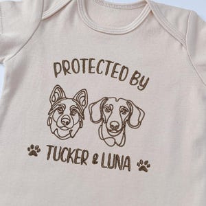 May include: A light beige short-sleeved bodysuit featuring brown embroidery of two dog faces, one resembling a German Shepherd and one a Dachshund. The text reads PROTECTED BY TUCKER & LUNA, flanked by two small brown paw print icons.