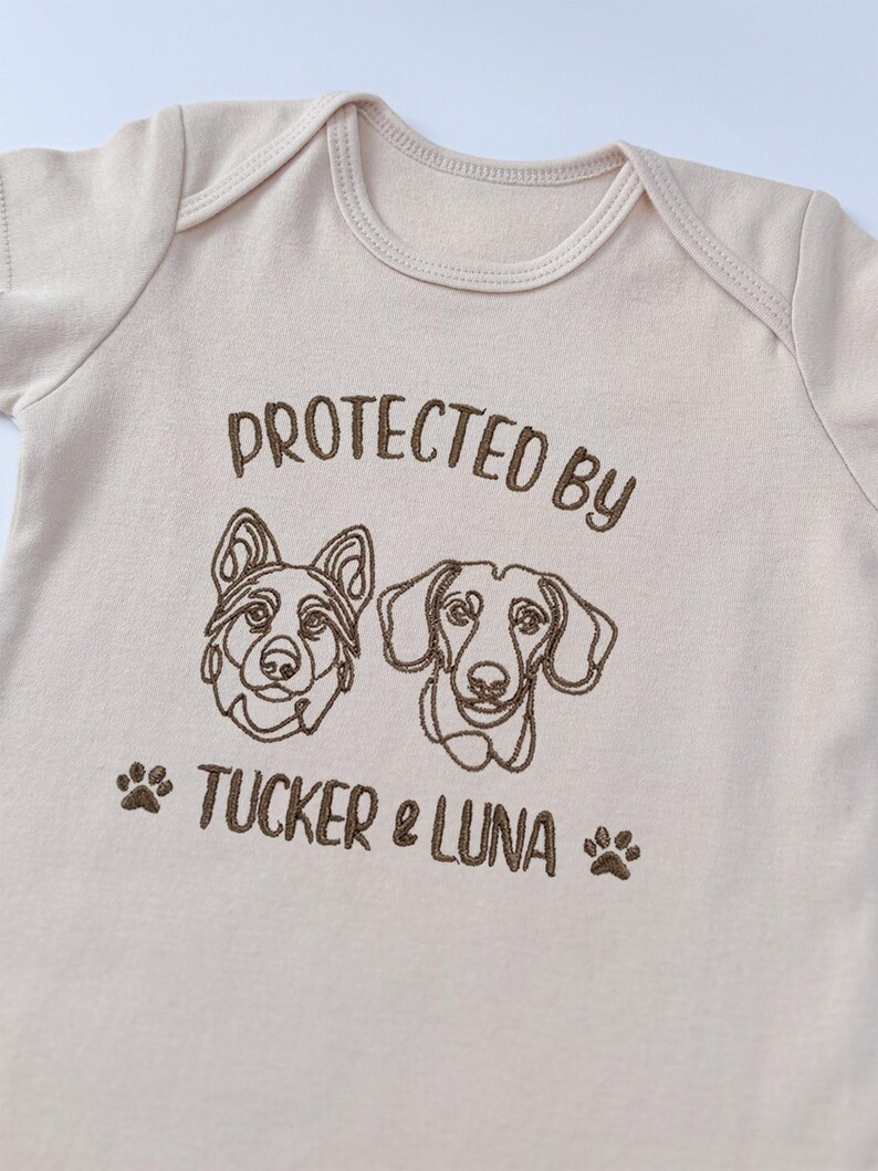 May include: A light beige short-sleeved bodysuit featuring brown embroidery of two dog faces, one resembling a German Shepherd and one a Dachshund. The text reads PROTECTED BY TUCKER & LUNA, flanked by two small brown paw print icons.