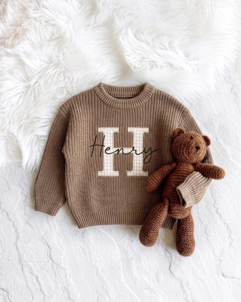 A cute neutral knit personalized toddler sweater designed for a baby shower gift, featuring custom applique initial embroidery and soft knit material on a white fur background.