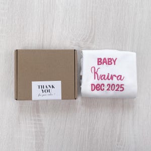 May include: A brown cardboard box with a white label reading THANK YOU for your order! next to a white fabric item featuring pink embroidered text that reads BABY Kaira DEC 2025. The items are arranged side by side on a light wood surface.