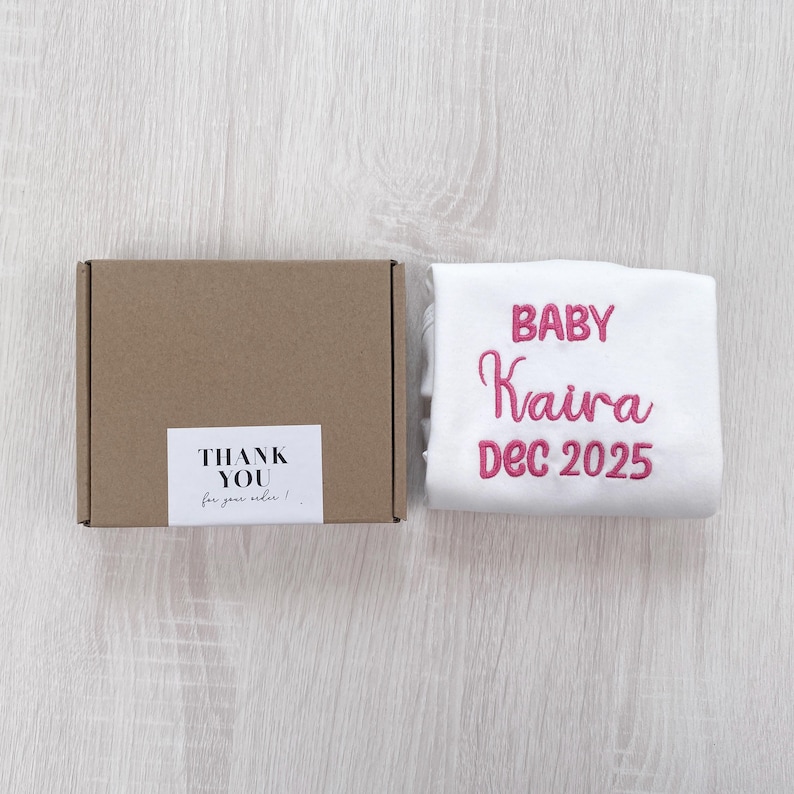 May include: A brown cardboard box with a white label reading THANK YOU for your order! next to a white fabric item featuring pink embroidered text that reads BABY Kaira DEC 2025. The items are arranged side by side on a light wood surface.