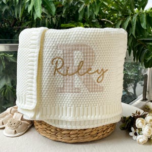 May include: A cream-coloured knitted baby blanket featuring a textured waffle pattern and a ribbed border. The centre displays a large, beige and white gingham-patterned letter R with the name Riley embroidered in gold cursive script.
