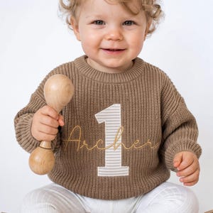 May include: A brown long-sleeve knit sweater featuring a large beige and white striped number one patch on the front with the name Archer embroidered in gold cursive script across the center of the number.