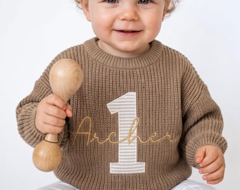 May include: A brown long-sleeve knit sweater featuring a large beige and white striped number one patch on the front with the name Archer embroidered in gold cursive script across the center of the number.
