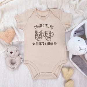 May include: A beige baby onesie with short sleeves. The onesie features the text "PROTECTED BY" above two dog illustrations and the text "TUCKER & LUNA" below, along with paw prints. The onesie is surrounded by soft toys.