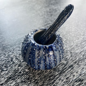 Pode incluir: Um conjunto artesanal de almofariz e pilão de cerâmica. O almofariz é azul escuro com detalhes prateados e uma superfície texturizada. O pilão é azul escuro e repousa dentro do almofariz. Ideal para moer ervas e especiarias.