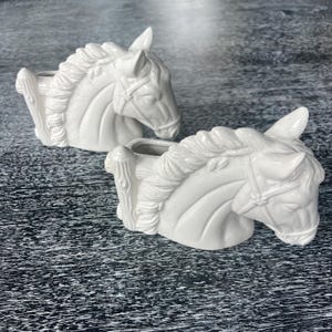 May include: Two white ceramic horse head planters. The horse heads are detailed with manes, bridles, and a wooden fence-like structure on the side. The planters are set against a dark, textured background.