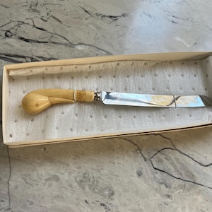 May include: A vintage butter knife with a light-colored handle and a silver blade, presented in a beige box. The handle has a curved design with a rounded end and a decorative band near the blade. The blade is rectangular with a slightly curved tip.