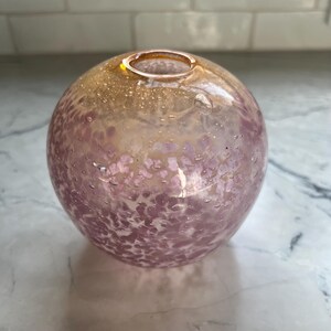 May include: A spherical glass vase with a speckled pattern in shades of pink and gold. The vase has a small opening at the top and is likely used for decorative purposes. The glass has a bubbly texture.