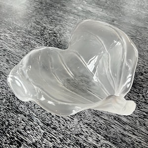 Puede incluir: Un cuenco de vidrio transparente en forma de hoja con un diseño texturizado y ondulado. El cuenco tiene un aspecto esmerilado y se encuentra sobre una superficie oscura y estampada. La forma del cuenco se asemeja a una cala.