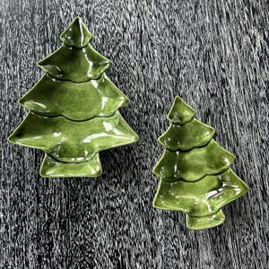 Peut inclure: Deux plats en céramique verts en forme de sapin de Noël. Les plats ont une finition brillante et sont conçus avec trois niveaux. Les plats sont sur une surface sombre et texturée. Idéal pour servir des collations ou comme pièces décoratives.
