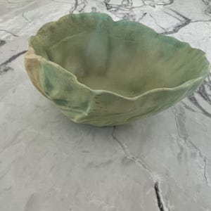 May include: A ceramic bowl with an organic, irregular shape and a wavy, ruffled rim. The exterior and interior surfaces feature a blended colour palette of soft sage green, pale yellow, and muted turquoise, creating a textured, layered appearance.