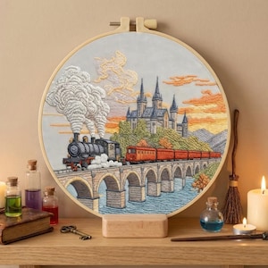 May include: Embroidered artwork in a wooden hoop depicts a train crossing a stone bridge towards a castle. The scene is set against a sunset sky with orange and yellow hues. The artwork is displayed on a wooden shelf with candles and potion bottles.