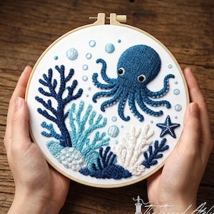 Peut inclure: Broderie sur tambour représentant un poulpe bleu, du corail et une étoile de mer. Le motif est réalisé sur fond blanc, avec des fils bleus et blancs. L'œuvre est tenue dans un cercle à broder en bois, mise en valeur par des mains.