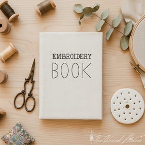 Può includere: Un libro di ricamo color crema con la scritta "EMBROIDERY BOOK" in nero. Il libro è circondato da forniture per ricamo, tra cui rocchetti di filo, forbici, un cuscino per aghi e un telaio, il tutto su una superficie di legno chiaro.