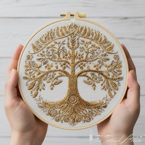 May include: A detailed gold-colored embroidered tree design on a white fabric, stretched within a wooden embroidery hoop. The tree features intricate branches, leaves, and roots. The artwork is held by hands, showcasing the craftsmanship and artistry of the piece.