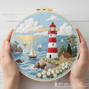 May include: Embroidered artwork in a wooden hoop depicting a coastal scene. A red and white striped lighthouse stands on a rocky shore, with sailboats on the blue water. White clouds and daisies complete the nautical design.