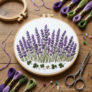 Op de afbeelding: Een geborduurde lavendel design in een houten borduurring, met paars en groen garen. De afbeelding toont borduur benodigdheden, waaronder garen, naalden, spelden en een schaar. De tekst "The Thread Atelier" is zichtbaar.