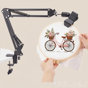 Puede incluir: Soporte de bastidor de bordado negro ajustable con abrazadera. Sostiene un bastidor de bordado redondo con un diseño de bicicleta rosa con cestas de flores. El bastidor de bordado está sostenido por las manos de una persona.