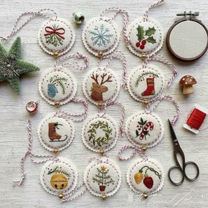 Pode incluir: Enfeites de Natal bordados com motivos festivos, incluindo um floco de neve e uma rena. Cada enfeite redondo tem uma borda ondulada e um pequeno sino dourado. Estão pendurados em barbante vermelho e branco.