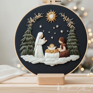 Peut inclure: Broderie d'une scène de Noël dans un cercle. L'œuvre représente une crèche avec des personnages, des arbres et une étoile. La palette de couleurs comprend du blanc, du marron, du vert et de l'or sur un fond bleu foncé. Le cercle est présenté sur un support en bois.