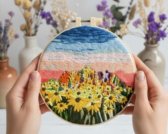 May include: A hand-stitched embroidery hoop featuring a landscape design. The top portion depicts a blue sky transitioning to a pink and peach sunset. The bottom half showcases a field of yellow and orange flowers with green foliage.