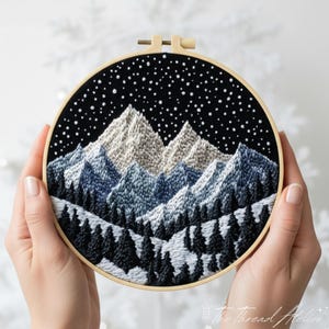 May include: Embroidered mountain landscape in a wooden hoop. The artwork depicts snow-covered peaks, dark blue and white trees, and a starry night sky. The embroidery is held against a blurred white background. The hoop measures approximately 20 cm in diameter.