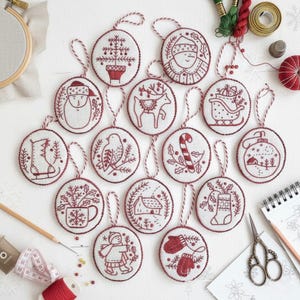 May include: A selection of embroidered Christmas decorations with red and white patterns. Each circular ornament displays a different festive illustration, such as a Christmas tree, Santa Claus, and a reindeer. The decorations are strung with twine and arranged on a white surface.