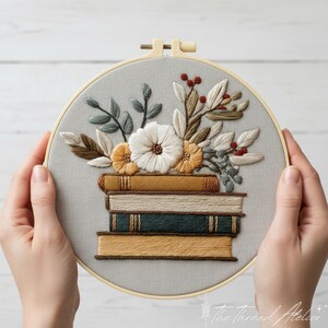 May include: Embroidered hoop art featuring a stack of books with floral accents. The design includes a stack of books in shades of brown and green, topped with white and yellow flowers, and various green and brown leaves. The embroidery is set against a gray fabric.