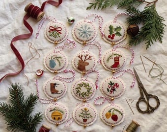 May include: Twelve circular fabric ornaments featuring hand-embroidered holiday motifs including mistletoe, snowflakes, holly, a reindeer, a stocking, an ice skate, a wreath, a candy cane, a jingle bell, a tree, and lights, with red and white string.