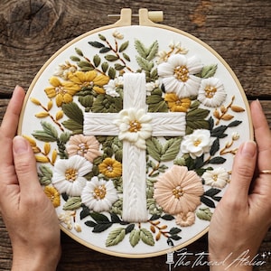Pode incluir: Um bastidor de bordado circular contendo tecido branco com um design bordado à mão de uma cruz branca central cercada por várias flores em tons de branco, amarelo e pêssego, acentuadas com folhas verdes. O texto The Thread Atelier é visível.