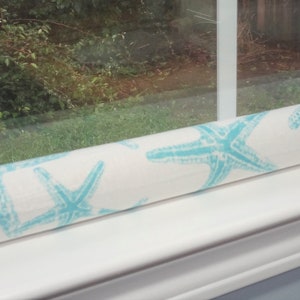 May include: White draft stopper with a blue starfish pattern. The draft stopper is rolled up and placed on a window sill.