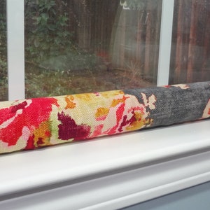 May include: A colorful floral patterned draft stopper, rolled up and placed on a window sill. The draft stopper is made of fabric and has a gray stripe running down the center.