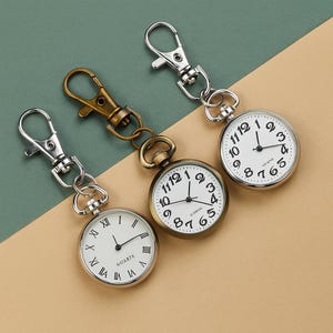 Könnte beinhalten: Drei Taschenuhren mit Schlüsselanhängern. Die Uhren haben weiße Zifferblätter mit schwarzen Ziffern und Zeigern. Die Uhrengehäuse sind silber- und bronzefarben. Auf einem Zifferblatt steht das Wort "QUARTE".