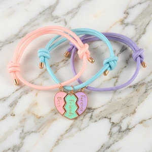 Pode incluir: Três pulseiras de amizade em tons pastel: rosa, azul e roxo. Cada pulseira tem um cordão com nós e detalhes em metal dourado. As pulseiras apresentam um pingente em forma de coração dividido em três seções, com as palavras "Best Friends Forever."