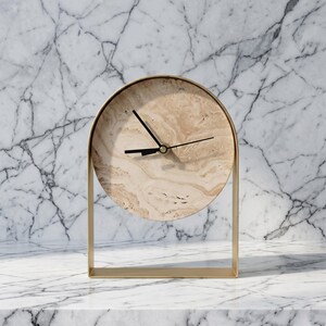 Op de afbeelding: Een moderne tafelklok met een ronde wijzerplaat en een goudkleurig metalen frame. De wijzerplaat heeft een beige marmerpatroon en de wijzers zijn zwart. De klok is ontworpen om op een vlakke ondergrond te staan.