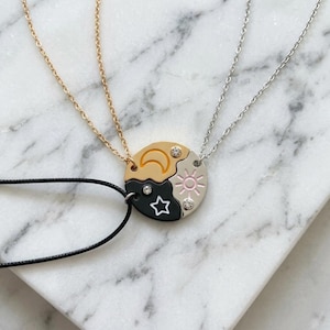 Peut inclure: Un ensemble de colliers avec un pendentif circulaire composé de trois pièces imbriquées. Une pièce dorée avec un croissant de lune, une noire avec une étoile et une argentée avec un soleil. Chaque pièce comporte un petit cristal clair.