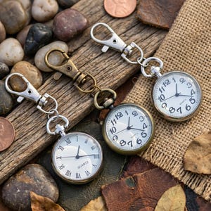 Puede incluir: Tres relojes de bolsillo con movimientos de cuarzo y cajas plateadas o bronceadas. Cada reloj tiene una esfera blanca con números y agujas negras. Los relojes están sujetos a llaveros con cierres.