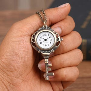 Op de afbeelding: Antieke sleutelhangerhorloge met een witte wijzerplaat en zwarte cijfers. Het horloge heeft een bronskleurige metalen behuizing en een bijpassende ketting. De sleutelvormige hanger heeft een decoratief ontwerp en een quartz uurwerk.
