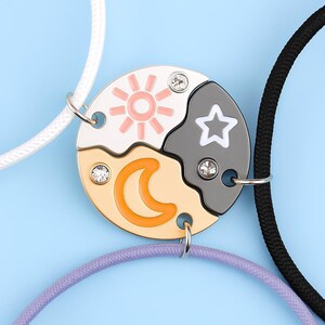 May include: A friendship necklace set featuring a sun, moon, and star design. The pendant is divided into three sections, each with a different celestial element. The sun is pink, the moon is orange, and the star is white. The necklace has a silver chain.