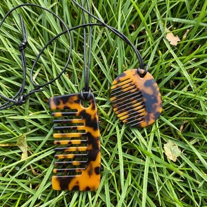 Puede incluir: Dos colgantes con forma de peine con estampado de carey sobre cordones negros, apoyados sobre hierba verde. Un colgante es rectangular, el otro ovalado. Los peines tienen dientes negros y están conectados a los cordones por un agujero en la parte superior.
