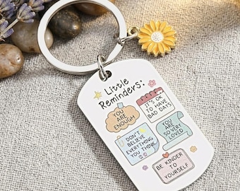 May include: A silver-colored keychain with a rectangular tag featuring the text "Little Reminders." The tag has colorful illustrations and phrases like "You are enough" and "Be kinder to yourself." A yellow sunflower charm hangs from the keychain.